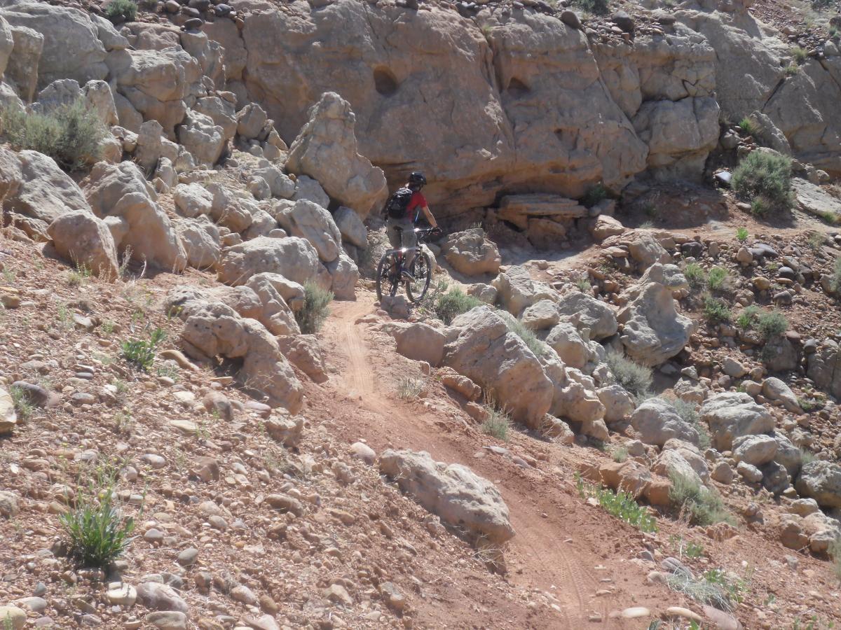 A mountain biker navigating a rocky trail surrounded by large boulders and sparse vegetation under a clear blue sky. Fire Sale mountain bike trail.