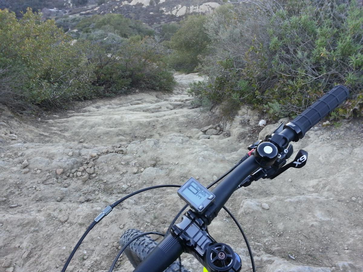 Close-up view of a mountain bike handlebar positioned on a rocky, uneven trail surrounded by shrubs and greenery, with a digital display showing speed and distance. The rugged terrain slopes downward, indicating a steep and challenging descent for cyclists. Aliso and Wood Canyons Wilderness Park mountain bike trail.