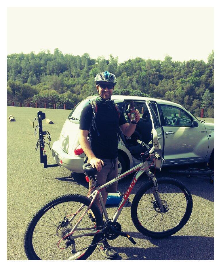 Giant Revel 1 29er: A person wearing a helmet and cycling gear stands next to a mountain bike, smiling and holding a beverage in one hand. In the background, a parked car is visible, along with a forested area. The scene appears to be a cycling or outdoor recreational spot.