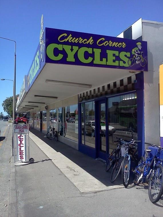 A storefront for "Church Corner Cycles," featuring a blue and green sign with a cyclist graphic. The shop front has large windows displaying the interior, and a sidewalk with several bicycles parked nearby. A sign placed outside the store also indicates the name of the shop. The scene is set under a clear blue sky.