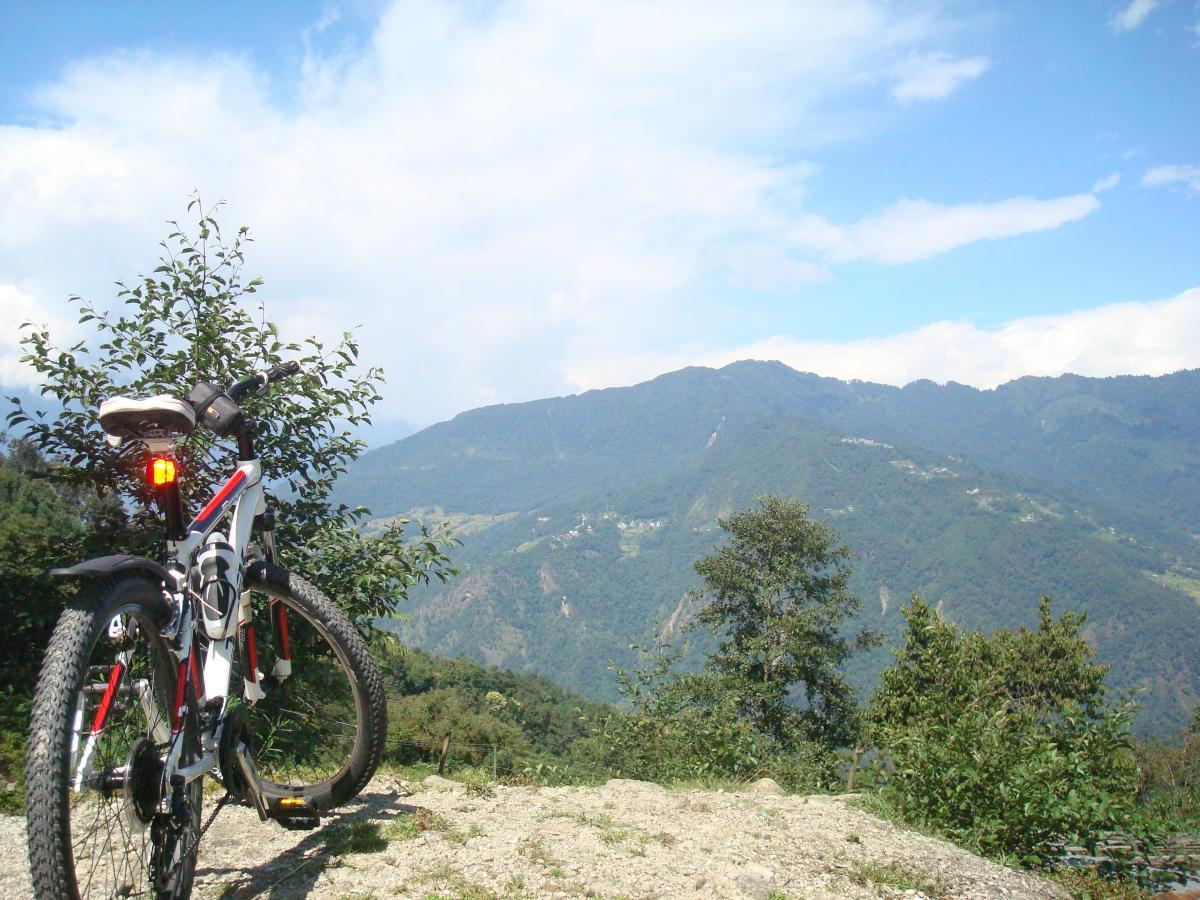 Trek 3700: A mountain bike is positioned on a gravel path overlooking a scenic valley with rolling green hills and distant mountains under a partly cloudy sky. Lush vegetation and trees frame the bike, creating a serene natural atmosphere.