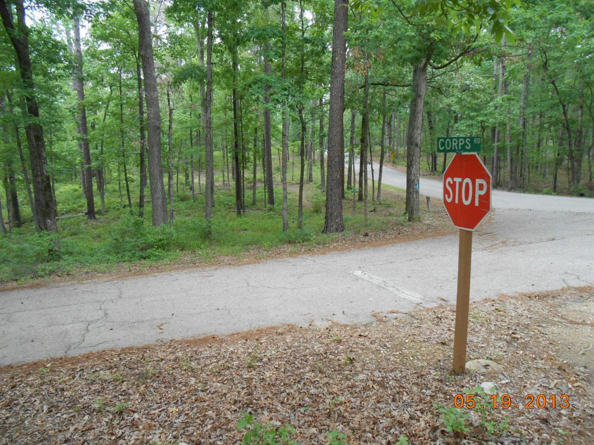 A dirt road in a wooded area with a "STOP" sign and a green street sign labeled "CORPS RD." Tall trees surround the area, and the path curves into the distance. The ground is covered with leaves and grass, creating a natural setting. Iron Mountain mountain bike trail.