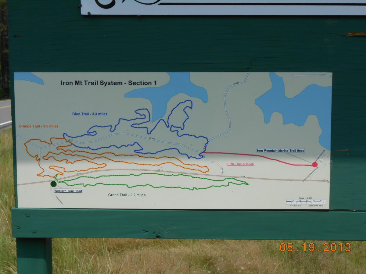 Map of the Iron Mountain Trail System, Section 1, displaying five marked trails: Orange Trail (3.5 miles), Blue Trail (3.3 miles), Pink Trail (5 miles), Green Trail (2.2 miles), and a connection to the Western Trail Head. The map includes a visual guide with trail lengths and locations relative to the Iron Mountain Marina Trail Head. Iron Mountain mountain bike trail.