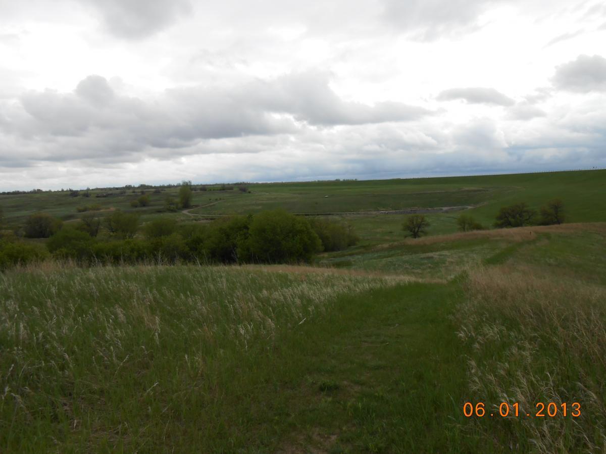 A scenic landscape featuring rolling green hills and a cloudy sky. The foreground shows a path winding through grassy terrain, surrounded by shrubs and trees. The background includes a distant view of more hills under a cloudy sky, suggesting an overcast day. The date at the bottom indicates it was taken on June 1, 2013. Pipestem Trail mountain bike trail.