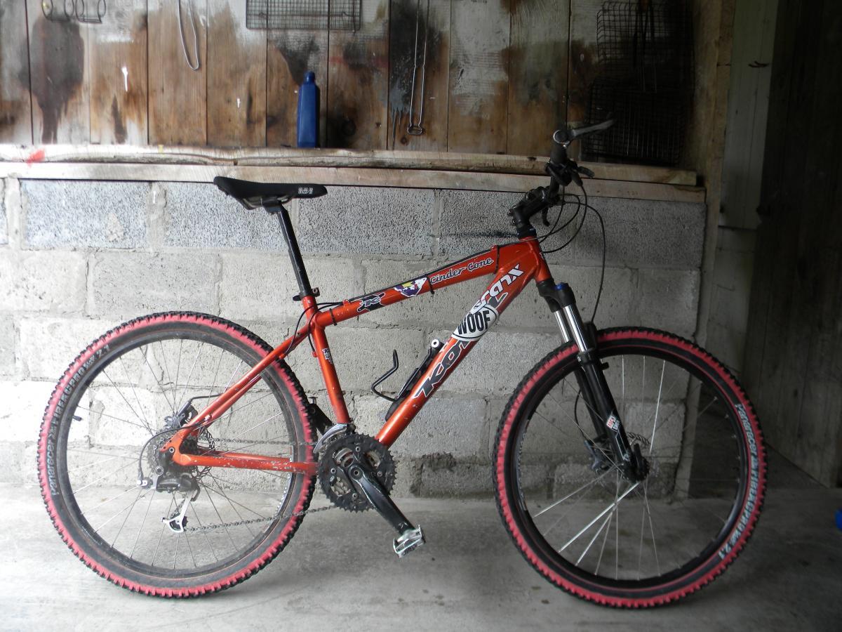 Kona Cinder Cone: An orange mountain bike with black and pink accents, leaning against a gray concrete wall in a garage or shed. The bike features a front suspension fork and multiple gears, with visible wear on the tires. Behind the bike, there is a wooden wall with various items hanging and a blue bottle on a shelf.