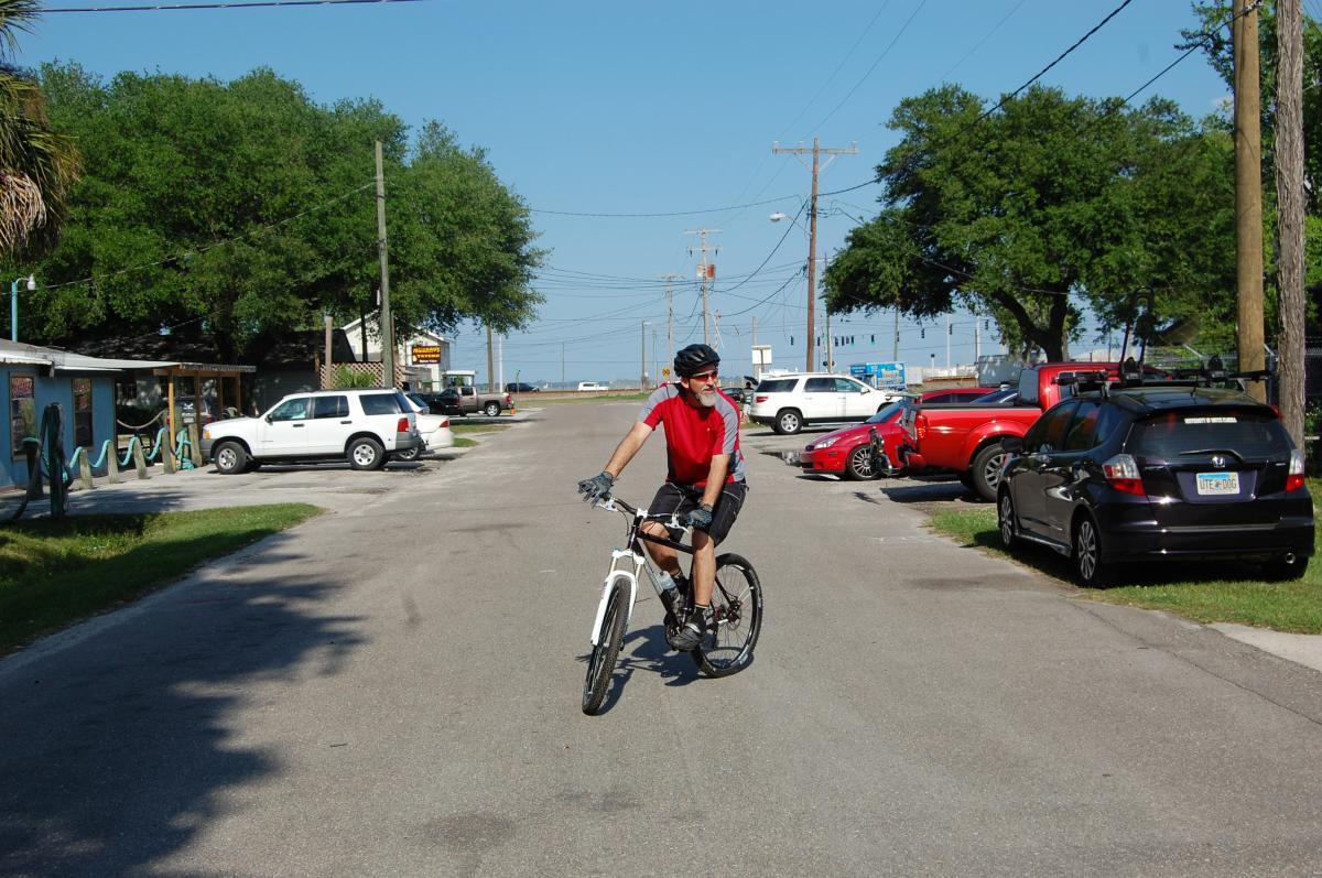 A cyclist in a red and gray jersey rides a white mountain bike down a residential street lined with parked cars. The background features greenery, utility poles, and a clear blue sky. Tillie Fowler Regional Park mountain bike trail.