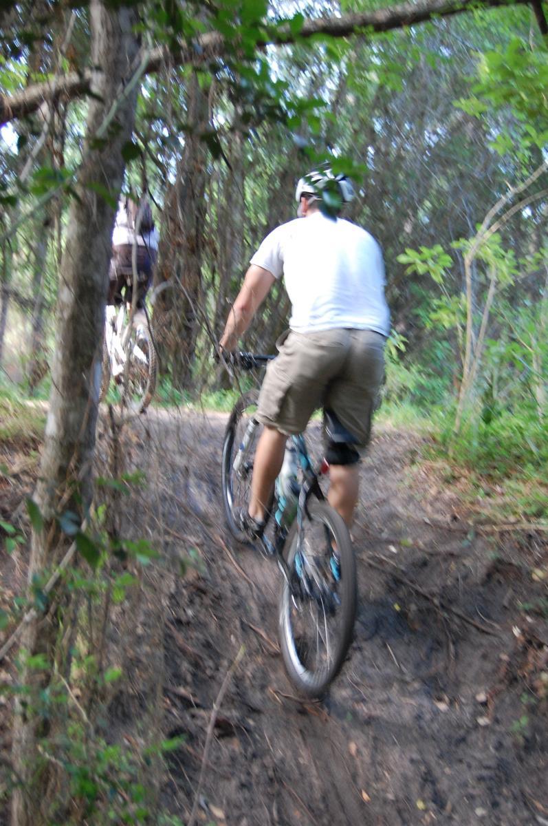 Two mountain bikers riding on a narrow dirt trail surrounded by dense greenery and trees. One biker is in the foreground, wearing a helmet and a white shirt, while the other is slightly behind, dressed in darker clothing. The trail appears rugged, with patches of dirt and foliage visible. Tillie Fowler Regional Park mountain bike trail.