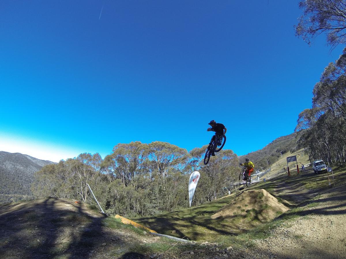 Two mountain bikers jumping off a dirt ramp in a scenic outdoor setting with blue skies and distant hills. One rider is in mid-air performing a trick, while the second rider is close to the ground preparing to take off. Surrounding trees and a white flag in the background emphasize the natural environment. Cannonball mountain bike trail.