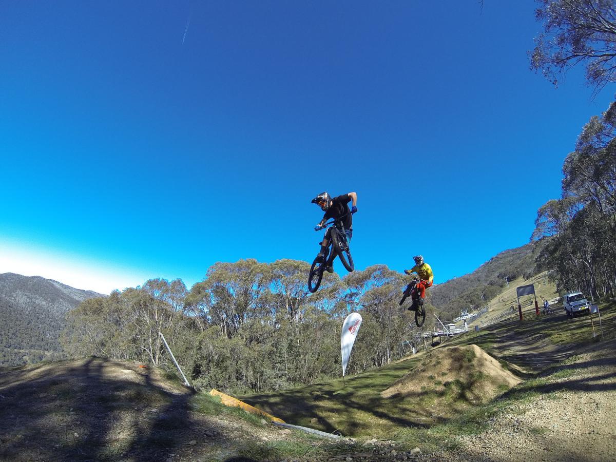 Two mountain bikers performing tricks in mid-air over dirt jumps, surrounded by trees and a clear blue sky. The scene captures the excitement of outdoor biking against a backdrop of hills and distant structures. Cannonball mountain bike trail.