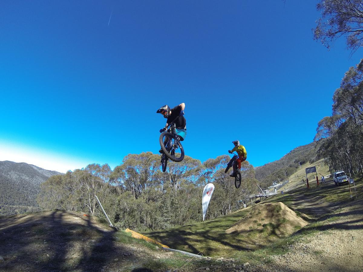 Two mountain bikers performing jumps over dirt mounds, set against a clear blue sky and green hills in the background. Cannonball mountain bike trail.