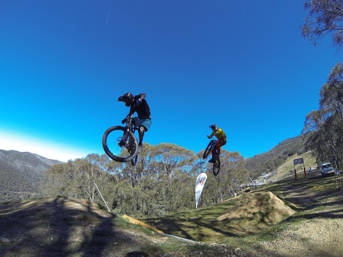 Two mountain bikers performing jumps on a dirt track surrounded by trees and mountains under a clear blue sky. The first biker, wearing a black shirt and helmet, is mid-air, while the second biker, dressed in a yellow shirt and orange pants, is also airborne. In the background, there are flags and a vehicle, indicating a biking event. Cannonball mountain bike trail.