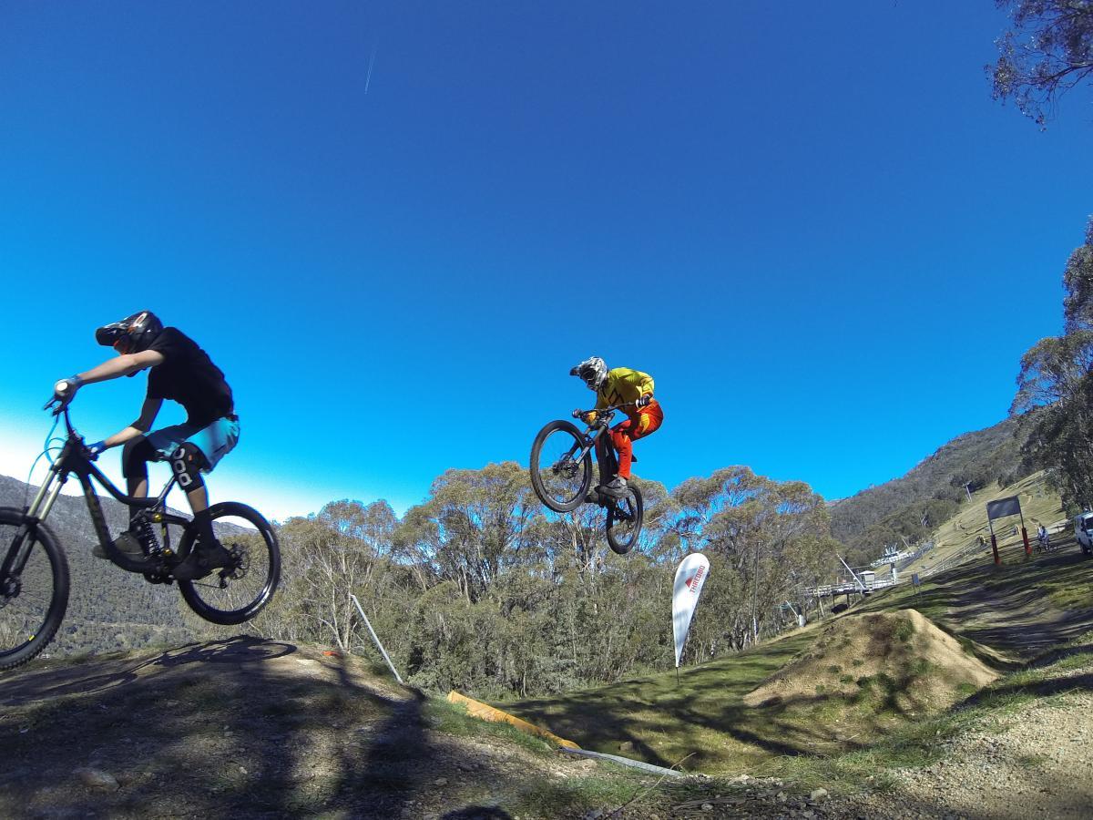 Two mountain bikers are airborne over a dirt jump in a scenic outdoor setting. One rider, dressed in a black shirt and blue shorts, is in the foreground, while the other, wearing a yellow shirt and orange pants, performs a jump in the background. The sky is clear and blue, with trees and a mountain slope visible in the backdrop, showcasing a vibrant day for biking. Cannonball mountain bike trail.