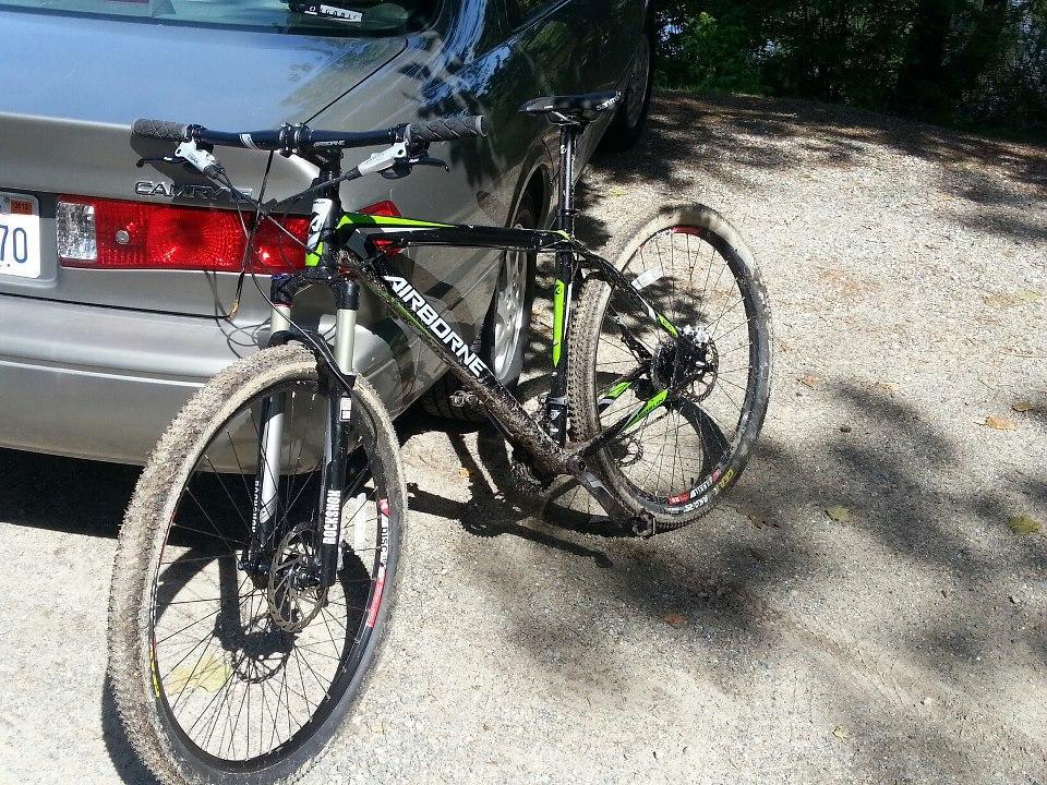 Airborne Goblin: A mountain bike with a black and green frame parked next to a gray car on a gravel surface, showing dirt on the tires and frame, indicating recent use.