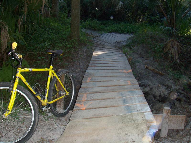 A yellow bicycle parked next to a wooden bridge leading into a wooded area. The path is surrounded by greenery, including tall palms and underbrush, indicating a natural setting. The bridge is made of wooden planks and extends over a sandy area. Soldier Creek Park mountain bike trail.