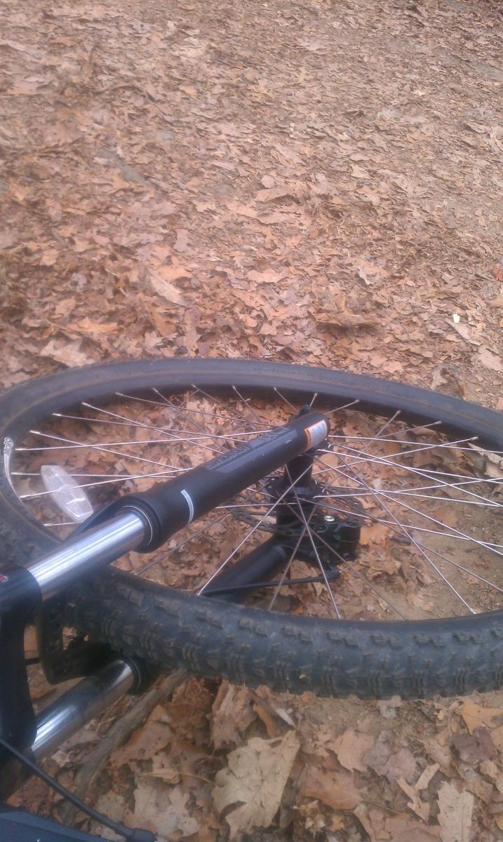 GT Karakoram: A close-up view of a bicycle lying on a ground covered with dry leaves. The focus is on the bike's front wheel and fork, with the wheel partially obscured by the leaf litter surrounding it.