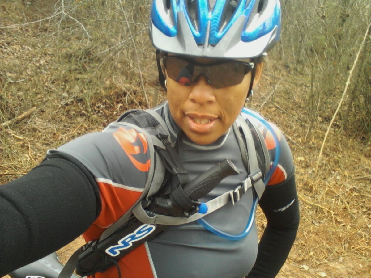 GT Karakoram: A person wearing a helmet and sunglasses, dressed in a cycling outfit, smiles at the camera while standing on a dirt trail surrounded by sparse vegetation. They are equipped with a hydration pack for biking.