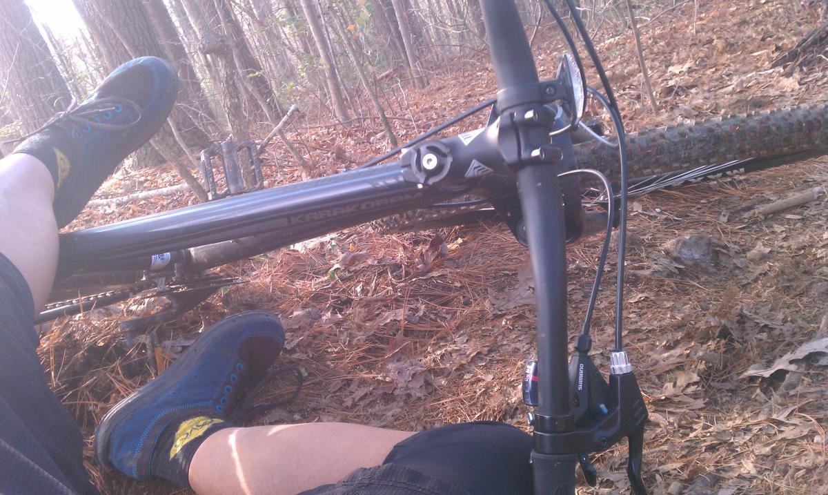 GT Karakoram: A person's legs are visible alongside a mountain bike lying on its side in a forested area covered with pine needles and leaves. The individual's feet are wearing blue shoes and black socks, with one leg resting on the bike frame. The background features trees and underbrush, suggesting an outdoor biking environment.