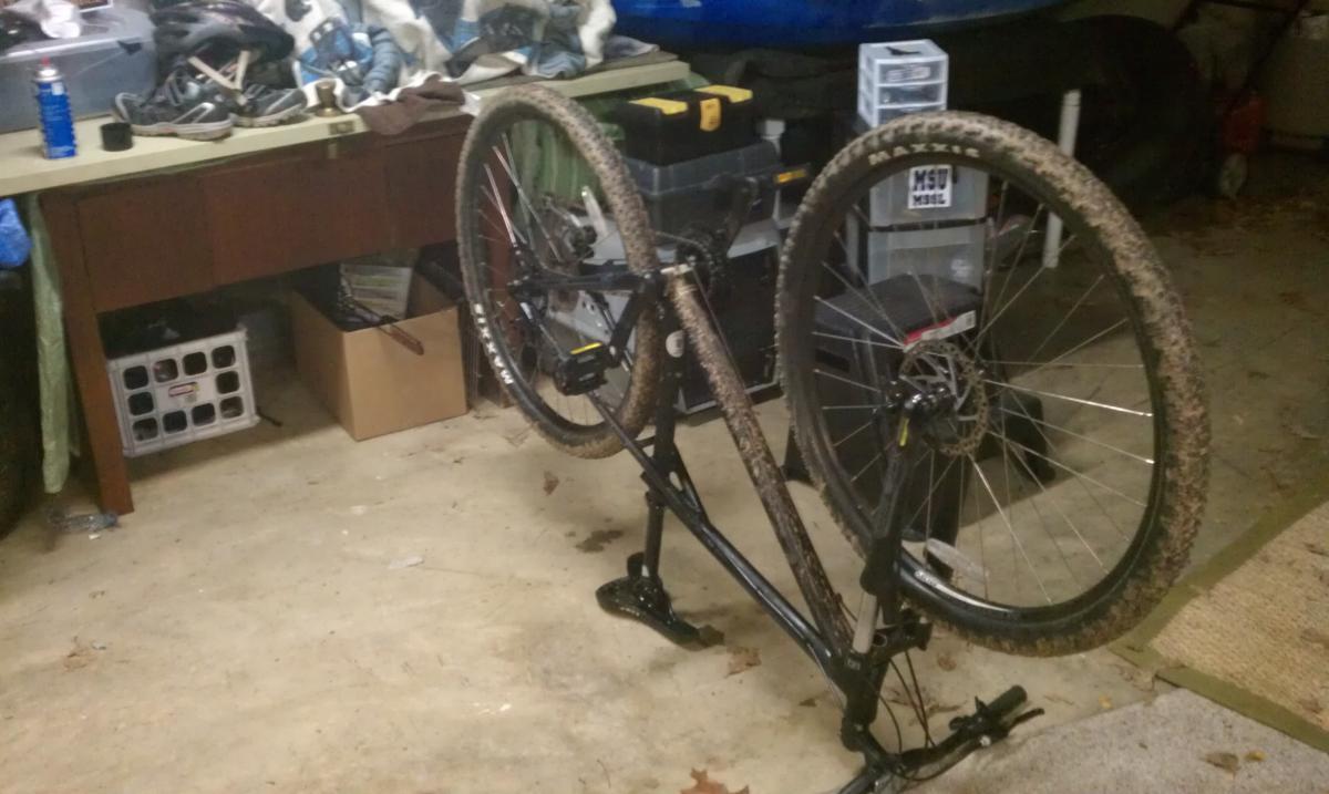 GT Karakoram: A mountain bike is shown upside down in a garage, with its wheels off the ground. The bike is covered in dirt, indicating recent use. Various items are scattered in the background, including a stack of helmets and a blue container. The floor is made of concrete, and there are boxes and tools visible in the cluttered space.