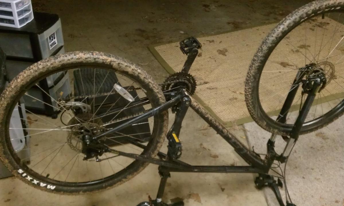 GT Karakoram: A black mountain bike resting upside down, showcasing its muddy tires and drivetrain, set against a garage floor with some scattered debris and storage boxes in the background.