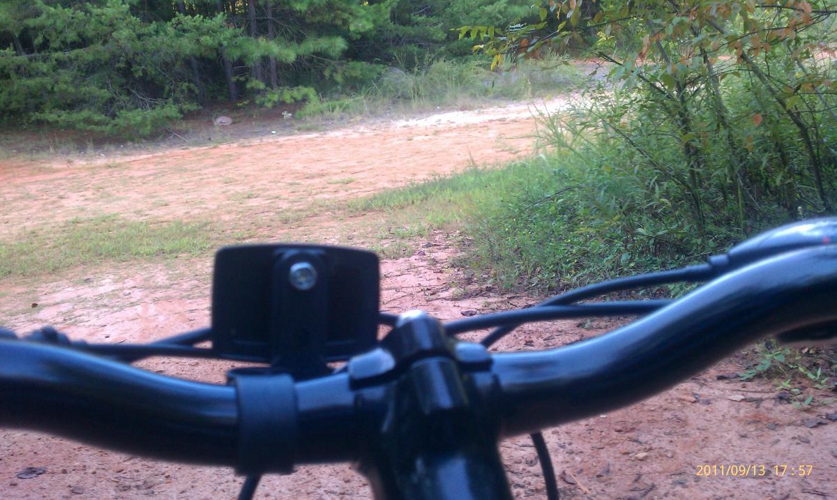 GT Karakoram: Close-up view of a bicycle's handlebars with a camera attached, overlooking a dirt path surrounded by greenery. The image captures the perspective of someone preparing to ride, with the path visible in front leading into a natural area.