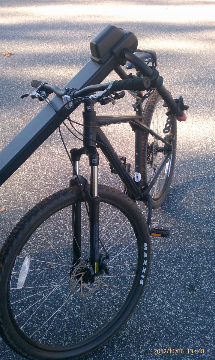 GT Karakoram: A close-up view of a mountain bike parked at an outdoor location. The bike features a black frame, thick tires with visible tread patterns, and a disc brake on the front wheel. The handlebars include a small black accessory mounted on them. The ground is paved, and there are leaves scattered around, indicating a natural setting.