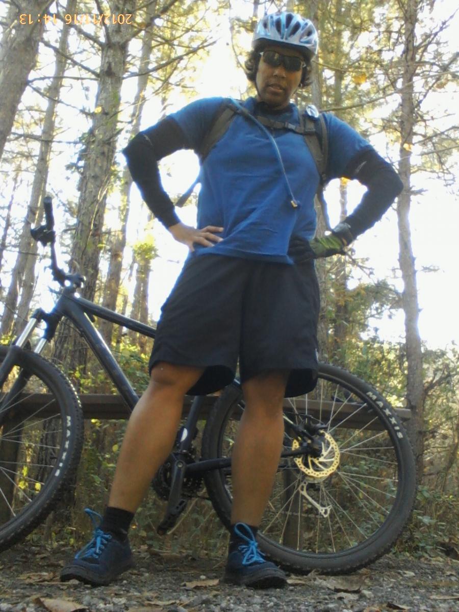 GT Karakoram: A cyclist standing confidently in a wooded area, wearing a blue t-shirt, black shorts, and a bike helmet. The person has sunglasses and is posing with hands on hips next to a mountain bike. Trees and foliage are visible in the background, creating a natural outdoor setting.