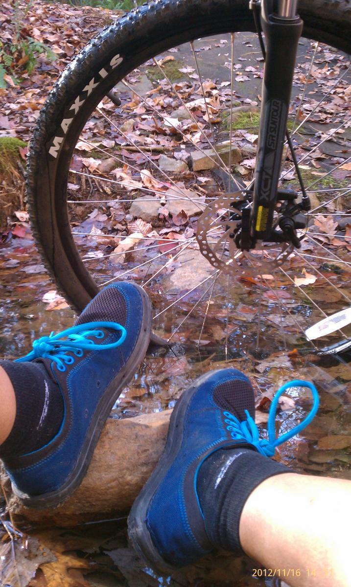 GT Karakoram: A pair of blue and black shoes resting on a rock in a shallow stream, with a mountain bike wheel partially visible in the background. The ground is covered with fallen leaves, indicating a natural outdoor setting.