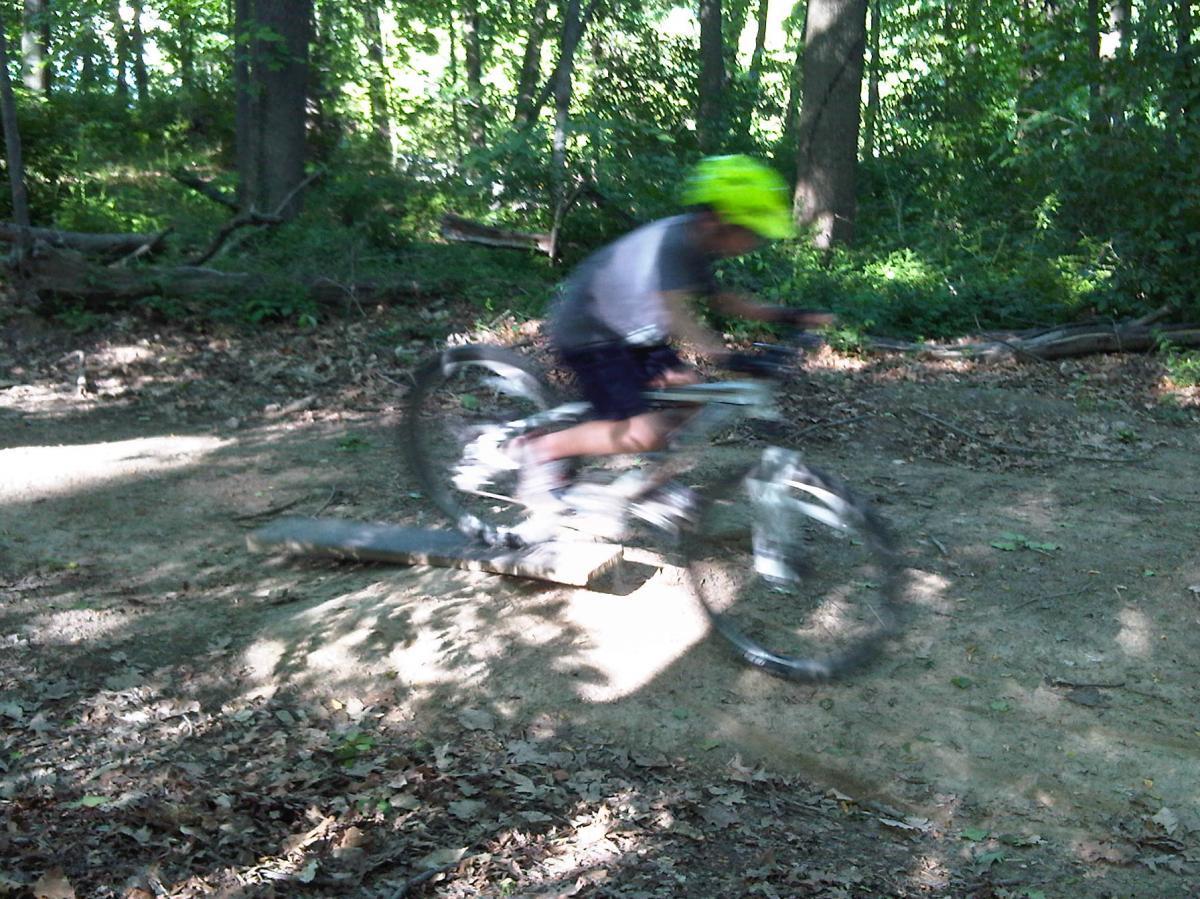 A blurred image of a person wearing a bright yellow helmet riding a mountain bike on a dirt trail in a wooded area, surrounded by trees and fallen leaves. Loughborough mill mountain bike trail.