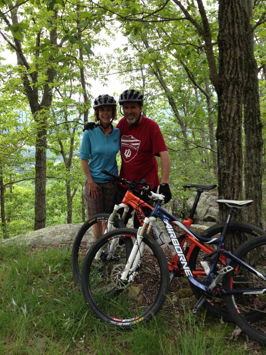 Airborne Hobgoblin: A couple poses for a photo while mountain biking in a lush green forest. They both wear helmets and sport casual biking attire. Two bicycles are leaning against a rock, surrounded by vibrant foliage and trees.