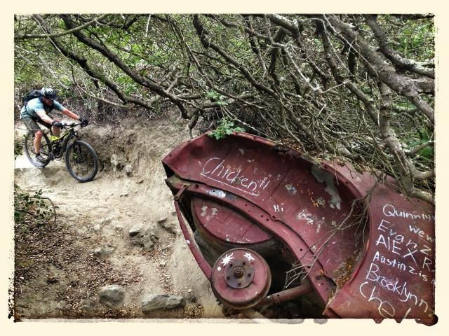 A mountain biker navigates a narrow, rocky trail surrounded by dense foliage, while an old, rusty piece of metal with graffiti is partially visible in the foreground. The scene captures the contrast between nature and remnants of human presence in an outdoor recreational area. Aliso and Wood Canyons Wilderness Park mountain bike trail.