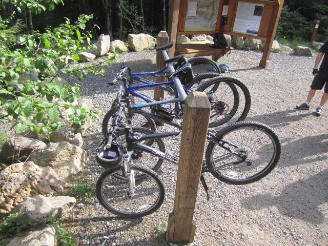 Four mountain bikes are secured to a wooden post in a gravel area surrounded by trees. A map board can be seen in the background, providing information about the trail or location. A person can be seen nearby, dressed in cycling gear. Sandy Ridge mountain bike trail.