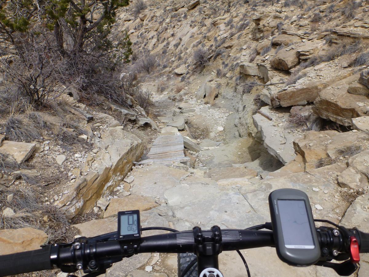 A mountain bike’s handlebars and digital display are visible in the foreground, facing a rocky and uneven trail that slopes downhill. The trail is bordered by sparse vegetation and dry terrain, with exposed stones and dirt creating a rugged landscape. The scene captures a challenging biking route in a natural setting. South Shore Lake Pueblo mountain bike trail.