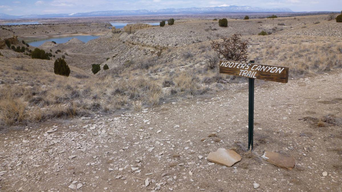 A wooden trail sign for "Hooter's Canyon Trail" is positioned on a rocky dirt path. The surrounding landscape features dry, grassy terrain and distant hills with a lake visible in the background under a cloudy sky. South Shore Lake Pueblo mountain bike trail.