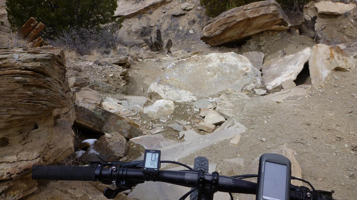 A view from the handlebars of a mountain bike, showing a rocky trail with large boulders and uneven terrain, surrounded by sparse vegetation and rugged terrain. The bike's display shows speed and other metrics, highlighting an adventurous outdoor setting for cycling. South Shore Lake Pueblo mountain bike trail.