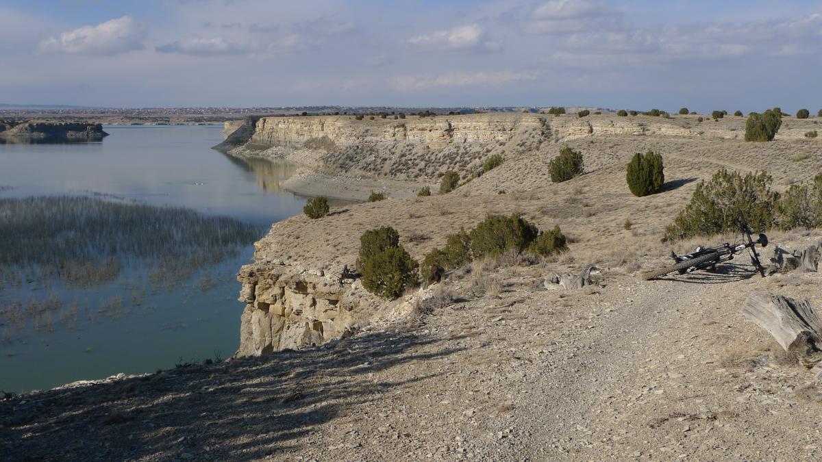 A scenic view of a rocky landscape overlooking a calm lake, with cliffs in the background. The foreground features a dirt path lined with sparse vegetation and a bicycle resting on the ground. The sky is partly cloudy, casting soft light over the tranquil scene. South Shore Lake Pueblo mountain bike trail.