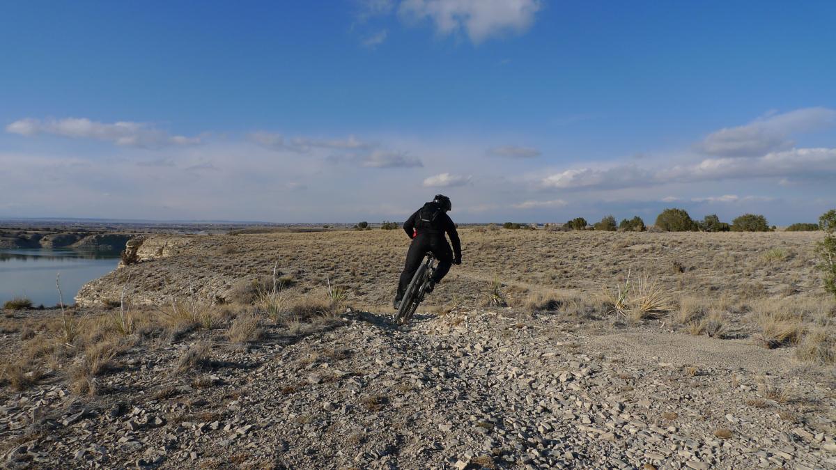 A mountain biker riding on a rocky trail along a lakeshore, with a vast landscape of dry grass and shrubs under a blue sky with scattered clouds. South Shore Lake Pueblo mountain bike trail.