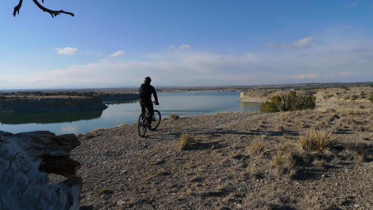 A cyclist wearing a helmet rides along a rocky path overlooking a calm lake surrounded by hills and sparse vegetation under a clear blue sky. South Shore Lake Pueblo mountain bike trail.