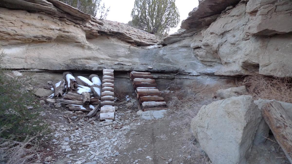 An abandoned area in a rocky landscape featuring a staircase made of wooden logs, leading up to a rocky overhang. Surrounding vegetation includes dry grass and sparse bushes, and there is some snow on the ground near the logs. The setting appears natural and rugged, suggesting a remote location. South Shore Lake Pueblo mountain bike trail.