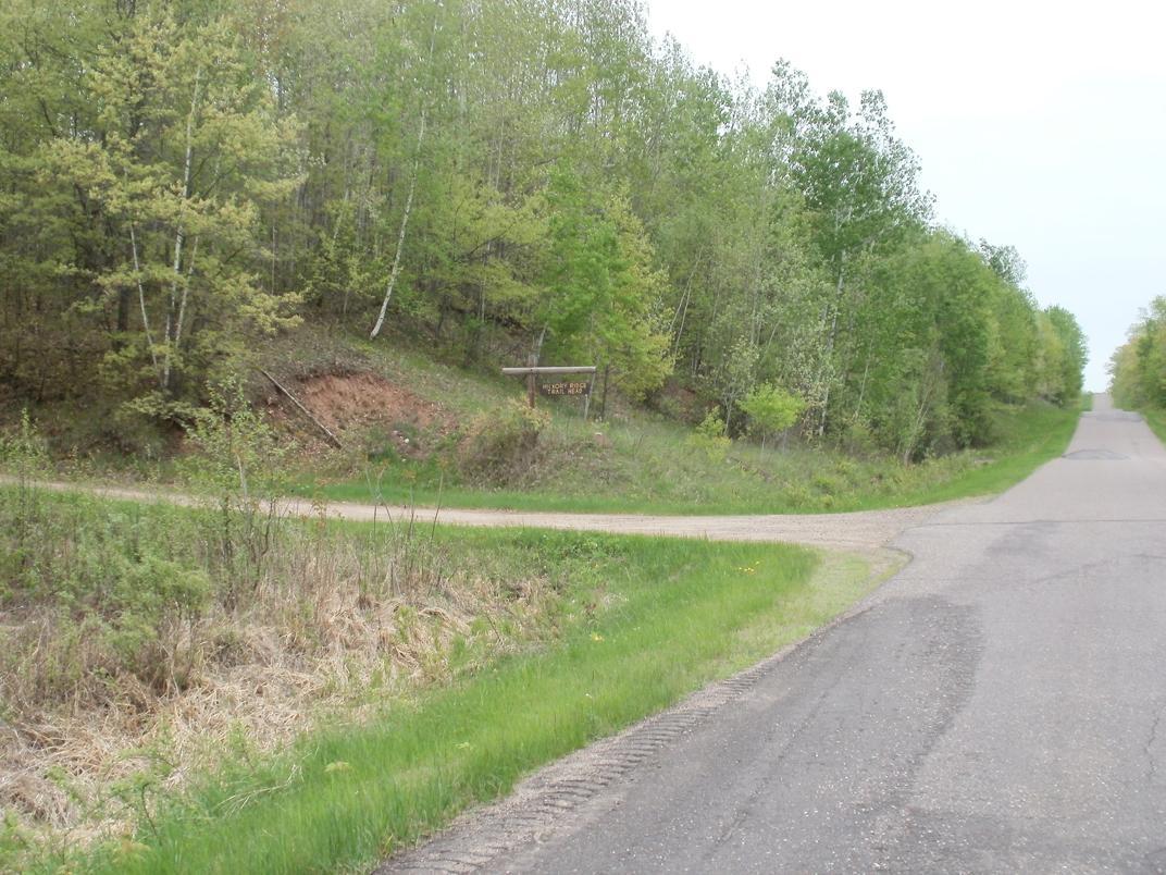 A rural scene depicting a split in a dirt road at the edge of a grassy area, surrounded by lush green trees. In the background, a sign is partially visible, indicating a turnoff. The scene is peaceful, with a clear sky above and new foliage suggestive of spring or early summer. Hickory Ridge mountain bike trail.
