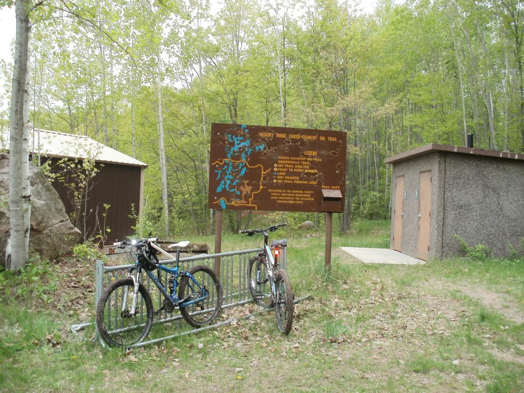 Bicycles parked at a bike rack near a trailhead, with a sign detailing the cross-country ski trail options. A small restroom building is visible next to the sign, surrounded by green trees and foliage in a natural setting. Hickory Ridge mountain bike trail.