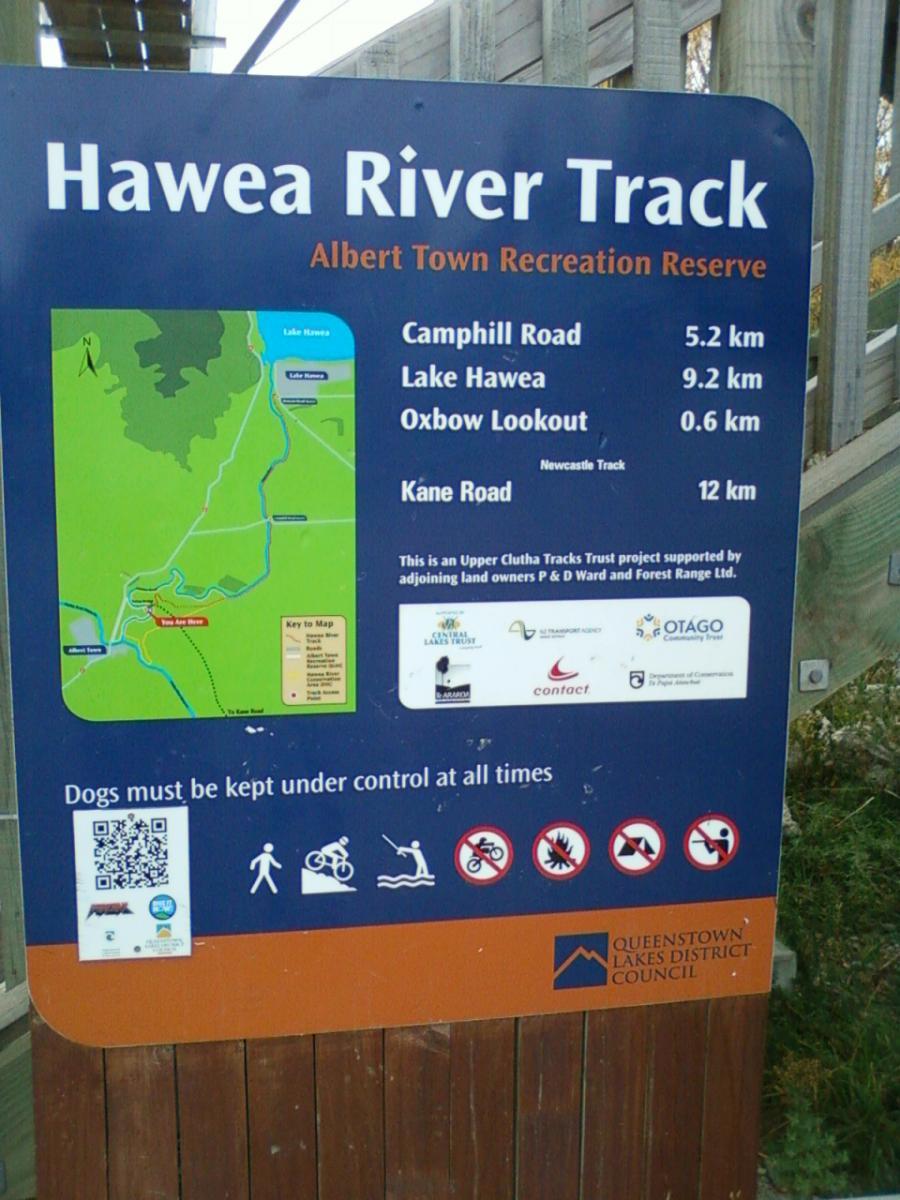 Sign for the Hawea River Track at Albert Town Recreation Reserve. The sign includes a map highlighting locations such as Camphill Road (5.2 km), Lake Hawea (9.2 km), Oxbow Lookout (0.6 km), and Kane Road (12 km). It contains information about track usage, safety regulations, and a QR code for additional details. Icons indicate activities allowed and prohibited, with a note that dogs must be kept under control at all times. Hawea River Track mountain bike trail.