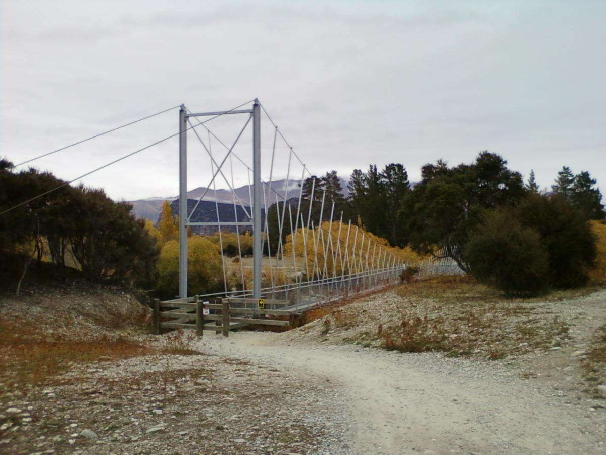 A suspension bridge with a metal frame crossing a dry riverbed, surrounded by trees and autumn foliage under a cloudy sky. A dirt path leads towards the bridge, which is supported by cables and features a gated entrance. Hawea River Track mountain bike trail.
