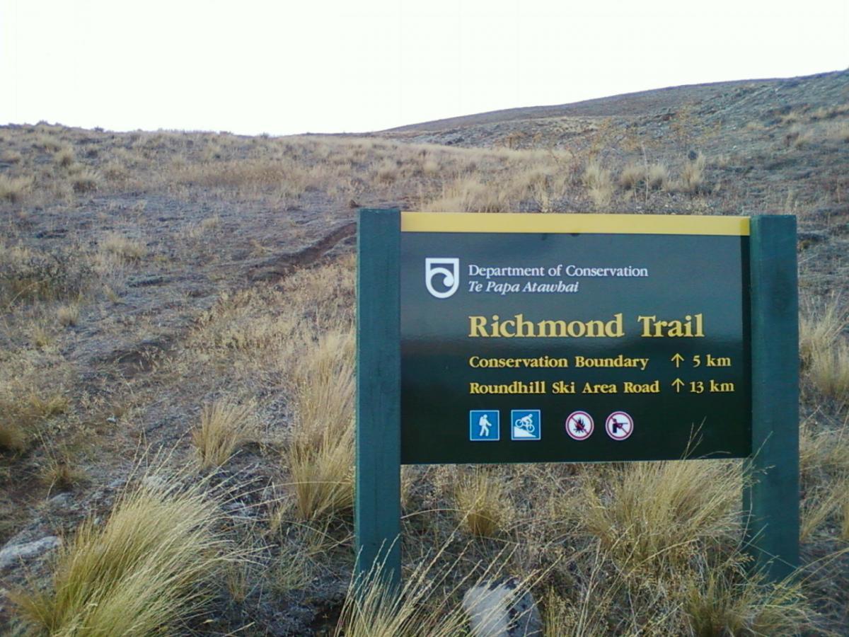 A sign for the Richmond Trail, indicating distances to the Conservation Boundary (5 km) and Roundhill Ski Area Road (13 km). The sign features icons for walking, biking, and wildlife, set against a backdrop of a grassy, hilly landscape. Richmond Trail Loop mountain bike trail.