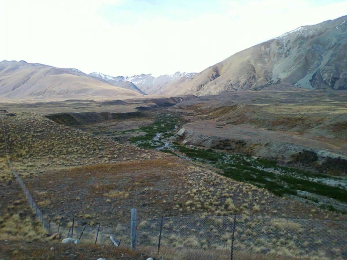 A scenic landscape featuring rolling hills and mountains in the background. The foreground showcases a dry riverbed surrounded by sparse vegetation and patches of green, indicating areas of life. A barbed-wire fence runs along the side, and the sky above is partly cloudy, creating a peaceful yet rugged natural environment. Richmond Trail Loop mountain bike trail.