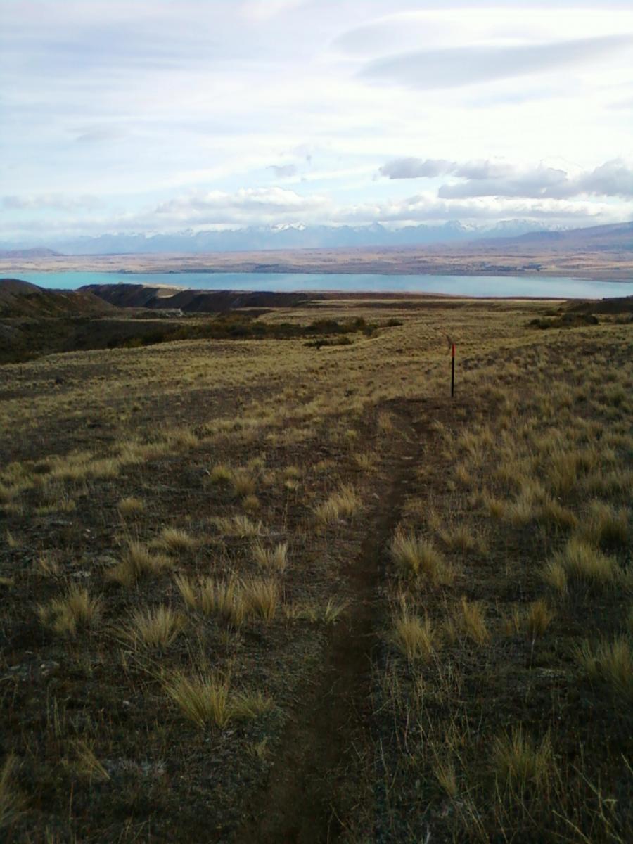 A scenic view of a grassy landscape with a visible dirt trail leading towards a body of water. In the background, mountains are partially covered by clouds, creating a tranquil and natural atmosphere. The foreground features patches of dry grass and small shrubs, while a trail marker is visible along the path. Richmond Trail Loop mountain bike trail.