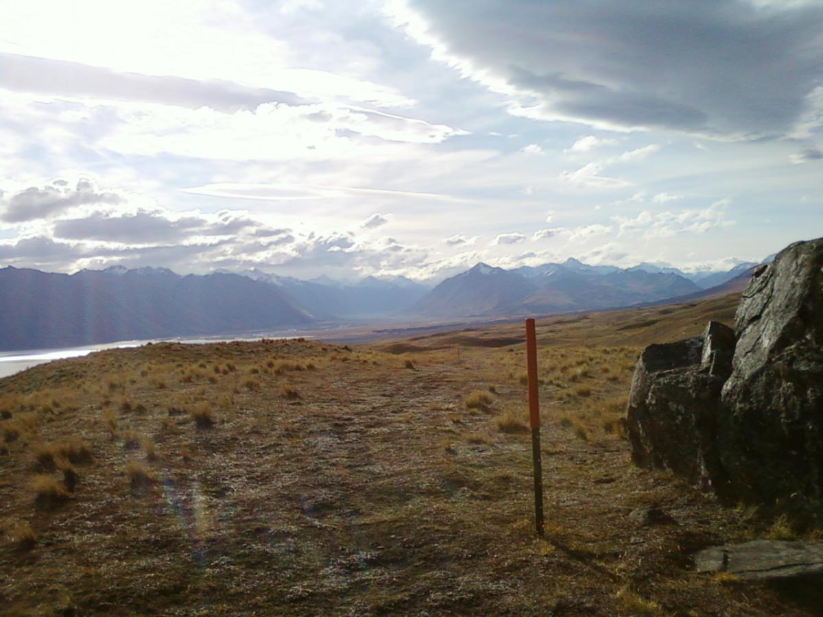 A scenic view of a mountainous landscape under a cloudy sky, featuring rolling hills covered in grass and a small orange post in the foreground. Snow-capped peaks can be seen in the distance, along with a body of water to the left. The atmosphere is serene and expansive, highlighting the natural beauty of the region. Richmond Trail Loop mountain bike trail.
