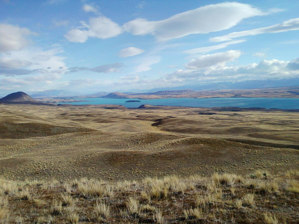 A panoramic view of a vast, rugged landscape featuring rolling hills and a tranquil blue lake. The sky is partly cloudy, with soft white clouds scattered across. In the distance, mountains are visible along the horizon, enhancing the natural beauty of the scene. The foreground consists of dry, golden grass and earthy tones, suggesting a remote and serene environment. Richmond Trail Loop mountain bike trail.