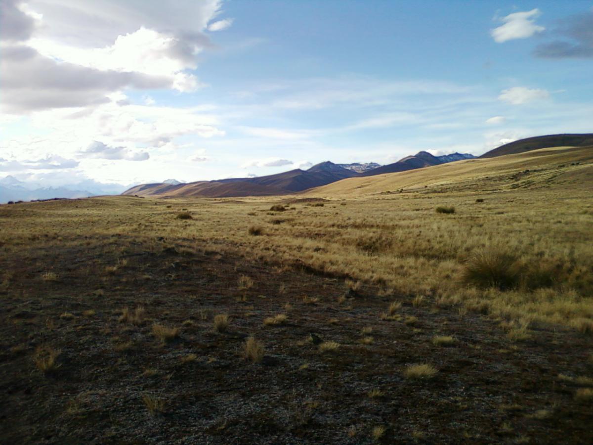 A panoramic view of a vast, grassy landscape under a partly cloudy sky, with rolling hills and distant mountains. The foreground features a mix of dry grass and rocky terrain, while the background displays snow-capped peaks silhouetted against the sky. Richmond Trail Loop mountain bike trail.