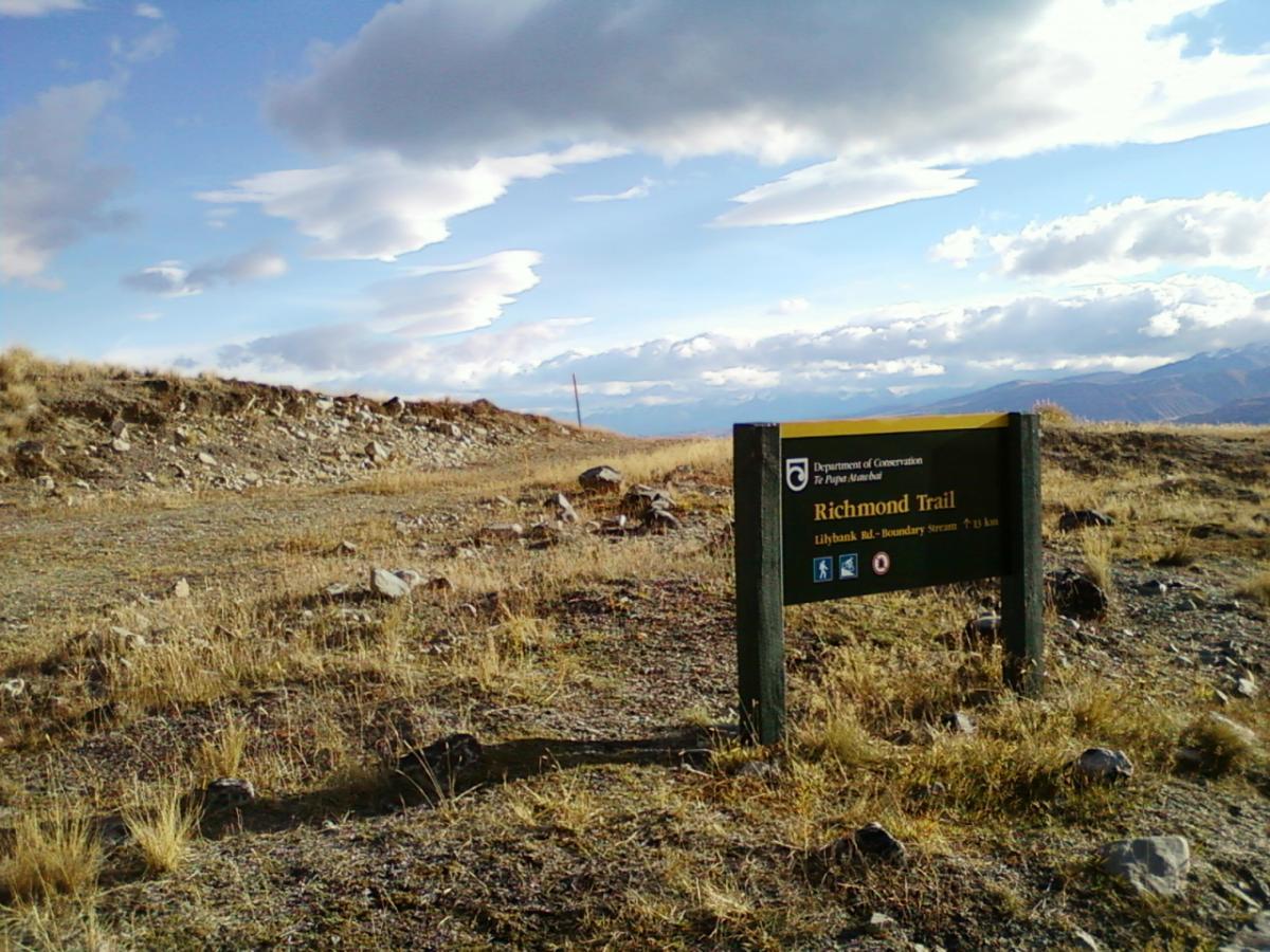 Sign marking the start of the Richmond Trail, set against a backdrop of rocky terrain and cloudy skies. The sign includes information about the trail length and nearby landmarks, with grassy and rocky ground surrounding it. Richmond Trail Loop mountain bike trail.