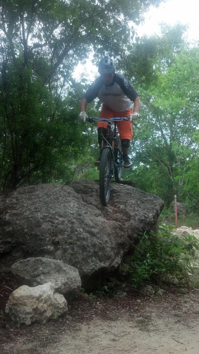 A person riding a mountain bike balances on a large rock in a wooded area. The cyclist is wearing a helmet, a short-sleeved shirt, and orange shorts, with protective gear on their knees. Lush greenery surrounds the scene, and a dirt path is visible in the background. Imba Red mountain bike trail.
